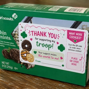 Pode incluir: Uma caixa retangular verde de biscoitos Girl Scouts Thin Mints com um peso líquido de 255 gramas. A embalagem apresenta um rótulo branco com o texto Thank you for supporting my troop, juntamente com ilustrações de vários tipos de biscoitos.