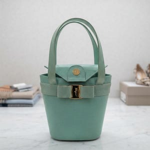 May include: A light mint green bucket bag with a textured lizard-skin pattern. The bag features two top handles, a fold-over flap with a gold-tone snap closure, and a decorative fabric bow across the front with a central gold-tone metal buckle.