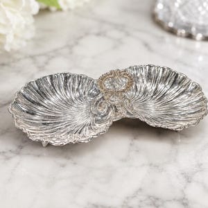 May include: A silver-toned metal serving dish featuring two conjoined shell-shaped bowls with a central decorative handle. The surface of the bowls has a ribbed, fan-like texture, and the edges are scalloped, creating a distinct dual-compartment design.