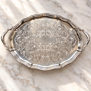 Peut inclure: Un plateau de service ovale en argent orné de motifs floraux et de volutes en relief sur toute sa surface. Le plateau présente un bord festonné et deux poignées incurvées sur les côtés, avec un centre ovale lisse et uni pour graver.