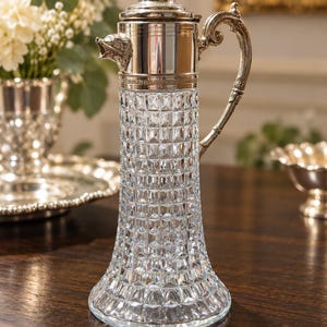 May include: A clear glass claret jug with a diamond-cut pattern, featuring a silver-plated metal collar, a hinged lid with a finial, and an ornate handle. The spout is adorned with a sculpted silver animal head, likely a boar or wolf.