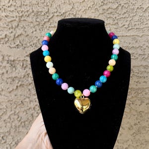 May include: A necklace featuring a strand of multicolored round beads in shades of pink, blue, yellow, green, and white, centered with a polished gold-tone heart-shaped pendant. The beads are arranged in a repeating pattern around the circular chain.