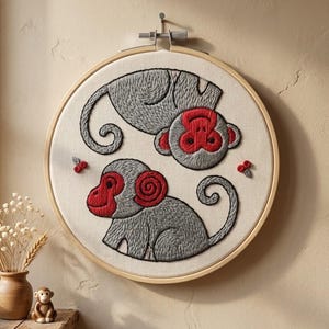 May include: A circular wooden embroidery hoop containing a cream-coloured fabric panel. The fabric features two embroidered monkeys with grey bodies and bright red faces and ears, positioned in a circular arrangement with small red floral accents.