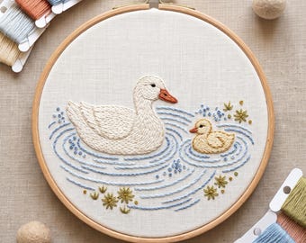 May include: A circular wooden embroidery hoop containing a white fabric panel with a hand-stitched design of a white adult duck and a small yellow duckling swimming in blue water ripples, surrounded by small green floral accents and embroidery floss.