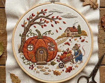 May include: An embroidery hoop containing a fabric panel with a stitched autumn scene. A large orange pumpkin house with a wooden door and window sits under a tree with red leaves. A mouse in blue overalls pushes a cart of pumpkins near a cottage.