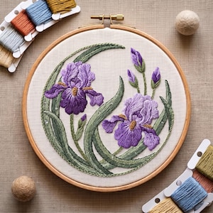 May include: A circular wooden embroidery hoop containing a fabric panel with a detailed needlepoint design of two purple iris flowers, green stems, and leaves. The embroidery features textured stitching in shades of violet, lavender, and green.