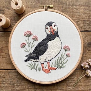 Könnte beinhalten: Ein runder hölzerner Stickrahmen mit weißem Stoff, auf dem ein Papageientaucher mit schwarzem, weißem und orangefarbenem Garn gestickt ist. Der Vogel ist von gestickten rosa Blumen und grünen Stängeln umgeben, daneben liegen zwei Holzspulen.