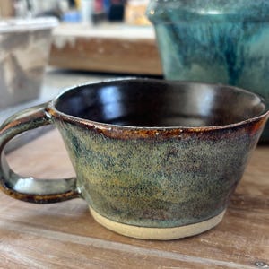 May include: A ceramic mug with a wide, flared rim and a sturdy handle. The exterior features a mottled glaze in shades of deep forest green, earthy brown, and hints of blue, while the base remains an unglazed, light beige clay colour.