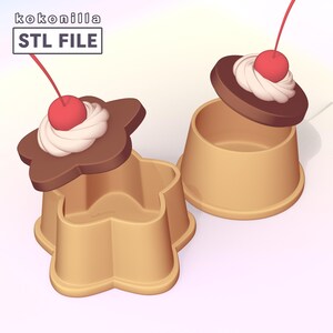 Op de afbeelding: Twee beige bakjes met deksels in de vorm van desserts met chocoladetopping. De ene is bloemvormig, de andere rond. Beide deksels hebben een witte toef slagroom en een rode kers. De tekst "kokonilla STL FILE" is duidelijk zichtbaar.