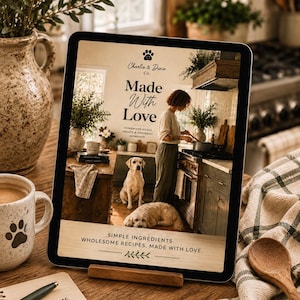 May include: A digital tablet displaying a cookbook cover titled Made With Love by Charlie & Dixie Co. The cover features a kitchen scene with a person, two dogs, and text reading Simple Ingredients. Wholesome Recipes. Made With Love.