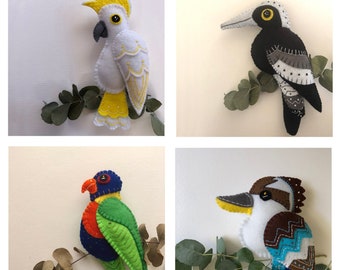 May include: Four felt bird ornaments, each with a different design and colour. The birds are perched on branches of eucalyptus leaves. The birds are a white cockatoo, a black and white bird with a yellow beak, a rainbow lorikeet, and a blue and brown kookaburra.