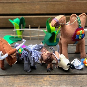 May include: A set of felt animal toys, including a brown cow, a gray donkey, a brown goat, a brown camel with a colorful saddle, a blue duck, and a yellow chick. The toys are arranged on a wooden surface.