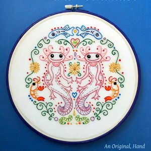 Puede incluir: Arte de bordado en un aro que representa dos ajolotes, un corazón, flores y camarones. El texto "The Inhabitants of Lake Xochimilco" está en la parte superior del aro. El texto "An Original, Hand Embroidery Pattern By Clara Batton Smith" está en la parte inferior del aro.