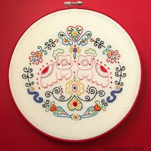 Op de afbeelding: Een witte borduurring met een kleurrijk handgestikt ontwerp van twee uilen met bloemen en krulletjes. De tekst "An Original, Hand Embroidery Pattern By Clara Batton Smith" staat onderaan de ring.