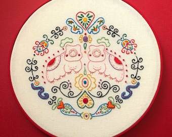 May include: A white embroidery hoop with a colorful hand-stitched design of two owls with flowers and swirls. The text "An Original, Hand Embroidery Pattern By Clara Batton Smith" is at the bottom of the hoop.