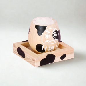 May include: A round wooden Daruma doll with a cow-print pattern of black spots on a light wood grain. The doll features a white painted face with a stern expression and the Japanese kanji for good fortune, and sits on a matching square wooden base.