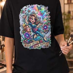 Pode incluir: Uma t-shirt preta com um gráfico colorido de uma pessoa a segurar um pincel de maquilhagem, rodeada por borboletas, batons e paletas. O texto "BLEND UNTIL MAGIC HAPPENS" está impresso numa faixa na parte inferior do desenho.