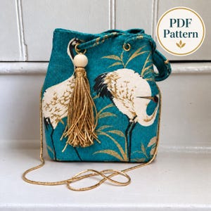 Puede incluir: Un bolso tipo cubo de color verde azulado con un patrón de grullas blancas y doradas, una correa de cadena dorada y una borla decorativa dorada con una cuenta de madera clara. El bolso incluye un gráfico circular en la esquina con el texto PDF Pattern.