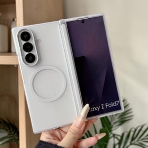 May include: A white protective phone case for a Samsung Galaxy Z Fold7, featuring a vertical slot for a stylus pen, a circular magnetic ring on the back, and a triple camera cutout. The screen displays the text Galaxy Z Fold7 in white font.