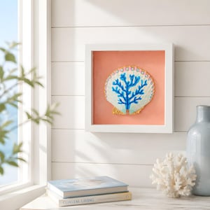 May include: A square white frame containing a large scallop shell painted with a bright blue coral design. The shell features gold-painted edges and small white dots along the top rim, set against a solid peach-colored background inside the frame.