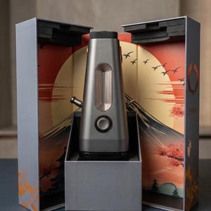 May include: A metallic gray, tapered electronic device with a circular dial and a vertical ribbed panel, housed in an open box featuring a printed illustration of a mountain, a large sun, and flying birds, with the text ENSO visible on the side.