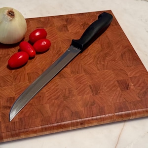May include: A square end-grain wooden cutting board with a visible grid pattern, featuring a stainless steel knife with a black handle, one whole white onion, and three small red cherry tomatoes placed on the surface.