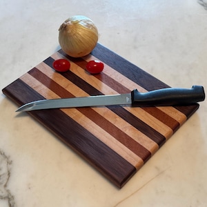 May include: A rectangular wooden cutting board featuring alternating stripes of dark walnut, cherry, and maple wood. A stainless steel knife with a black handle rests on the board, accompanied by a whole yellow onion and two small red tomatoes.