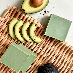 Op de afbeelding: Een lichtgroen rechthoekig stuk zeep en twee dunne plakjes groene zeep op een gevlochten rieten dienblad. Het dienblad bevat ook een gehalveerde avocado met pit, vier dunne plakjes avocado en één hele avocado met een donkere schil.