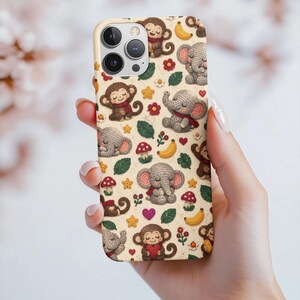 May include: A phone case featuring a repeating pattern of crocheted-style illustrations of monkeys and elephants, interspersed with small red mushrooms, yellow bananas, green leaves, yellow stars, and small pink and red hearts on a cream background.