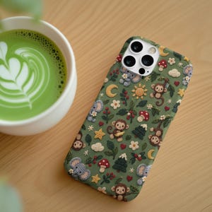 May include: A sage green smartphone case featuring a repeating pattern of crocheted-style motifs including brown monkeys, grey elephants, red mushrooms, yellow stars, crescent moons, white flowers, green pine trees, and small red hearts.