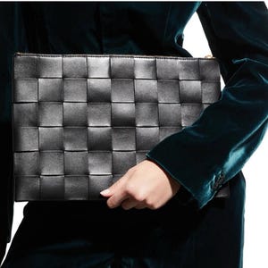 May include: A rectangular black clutch bag featuring a woven leather construction with a grid pattern. The bag has a smooth, semi-glossy finish and a gold-toned zipper closure along the top edge, providing a structured and uniform appearance.