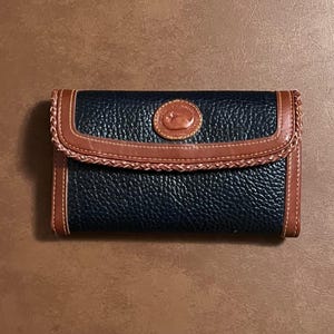 May include: A rectangular navy blue wallet with a textured leather surface, featuring tan leather trim with braided detailing along the flap edge. A circular tan leather patch with an embossed duck logo is centered on the front flap closure.