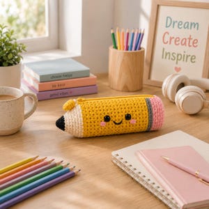 Puede incluir: Un estuche de ganchillo amarillo, negro y rosa con forma de lápiz y una cara sonriente. El estuche presenta una punta negra, un cuerpo amarillo, una banda gris y un extremo de goma rosa. Es un artículo suave y texturizado con cremallera.