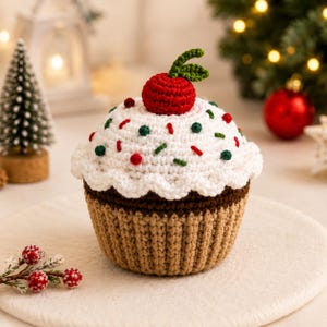 May include: A crocheted cupcake decoration featuring a tan base, white frosting, and a red cherry on top. The white frosting is adorned with small red and green sprinkles. The item is crafted with yarn and displays a textured, handmade appearance.