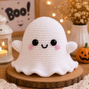 May include: A white crochet ghost figurine featuring a rounded head, two small arms, and a scalloped base. The ghost has two large black plastic eyes, a small embroidered black smile, and two circular pink stitched cheeks on its face.