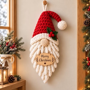 May include: A crocheted gnome wall hanging featuring a red hat with a white pom-pom, a beige nose, and a long white beard. A wooden sign hangs from the beard with the text Merry Christmas and a small heart icon. The gnome is suspended by a loop.