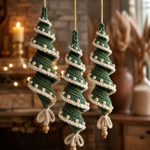 Könnte beinhalten: Drei hängende spiralförmige Weihnachtsbaum-Ornamente aus dunkelgrünem Garn mit weiß gehäkelten Rändern und kleinen weißen Perlen. Jedes Ornament hängt an einer geflochtenen Kordel und hat einen dekorativen Knoten sowie eine Holzperle am unteren Ende.