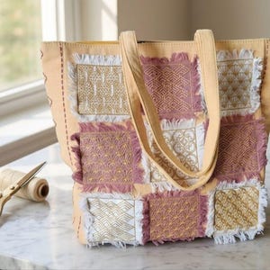 Peut inclure: Un sac fourre-tout beige orné d'un motif patchwork composé de carrés de tissu dans des tons rose, blanc et doré. Chaque panneau présente un motif géométrique brodé distinct avec des bords effilochés. Le sac possède deux anses assorties.