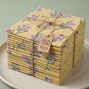 May include: A stack of 150 square napkins in a mustard yellow colour featuring a repeating floral pattern of pink flowers and green stems. The napkins are tied together with brown twine and include a small rectangular tag labelled 150 Pack.