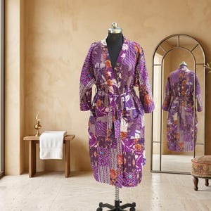 May include: A knee-length robe featuring a patchwork design in various shades of purple, accented with orange and white floral and geometric patterns. The garment includes long sleeves, a tie-waist belt, and two front pockets, displayed on a mannequin.