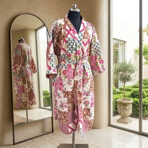 Puede incluir: Una bata de manga larga con un diseño de patchwork que presenta varios patrones florales y geométricos en tonos rosa, marrón y blanco. La prenda incluye un cuello tipo chal, un cinturón para anudar y una textura acolchada.