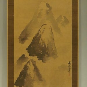May include: A vertical scroll painting showing a mountain landscape in shades of brown and grey. The artwork features three prominent mountain peaks and a small structure at the base, with Japanese calligraphy.