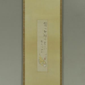 May include: A traditional Japanese hanging scroll with a cream-colored background. The scroll features a rectangular image with Japanese text and a small illustration. Green and gold decorative elements are present.