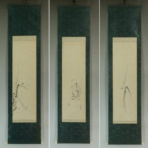Op de afbeelding: Drie verticale Japanse schilderijen op rol. Elk heeft een crèmekleurig middenpaneel met een zwarte inktillustratie. De illustraties tonen bamboe, een figuur en een plant. De panelen zijn omlijst door een decoratieve rand en een donkergroene stof.