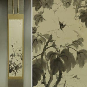 May include: A vertical hanging scroll featuring a monochromatic ink wash painting of a flowering plant with large blossoms and broad leaves. The artwork is mounted on a tan and grey fabric border with two thin hanging cords at the top edge.