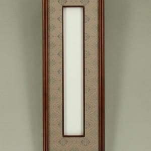 Puede incluir: Una obra de arte mural rectangular y vertical con un marco de madera marrón oscuro. El interior presenta una tela estampada en beige y gris, que enmarca un espacio rectangular blanco central. Se suspende con un alambre fino.