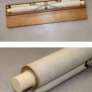 May include: A rolled scroll with ivory-colored end pieces, secured with a silver-colored chain, rests inside a wooden box. The scroll is cream-colored and appears to be made of paper or fabric. The box is made of light-colored wood.