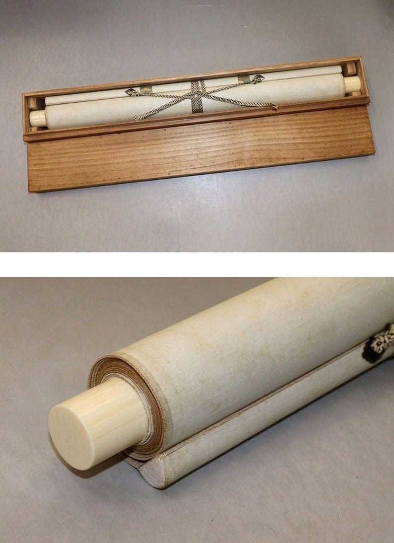 May include: A rolled scroll with ivory-colored end pieces, secured with a silver-colored chain, rests inside a wooden box. The scroll is cream-colored and appears to be made of paper or fabric. The box is made of light-colored wood.