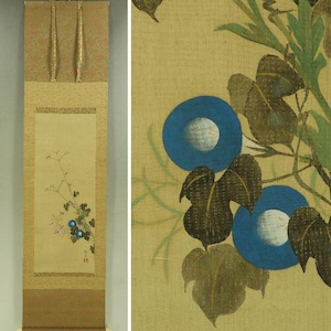 May include: A vertical hanging scroll featuring a painting on a light beige fabric background. The artwork depicts a botanical scene with green leaves, thin stems, small pink flowers, and two prominent blue morning glory blossoms with white centres.
