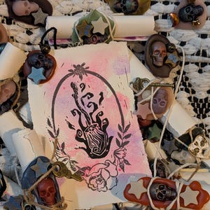 May include: A rectangular paper card with a black ink block print of a stylized flame inside an oval frame, set against a pink and purple watercolor background. The card is surrounded by multiple rolled paper scrolls sealed with wax.